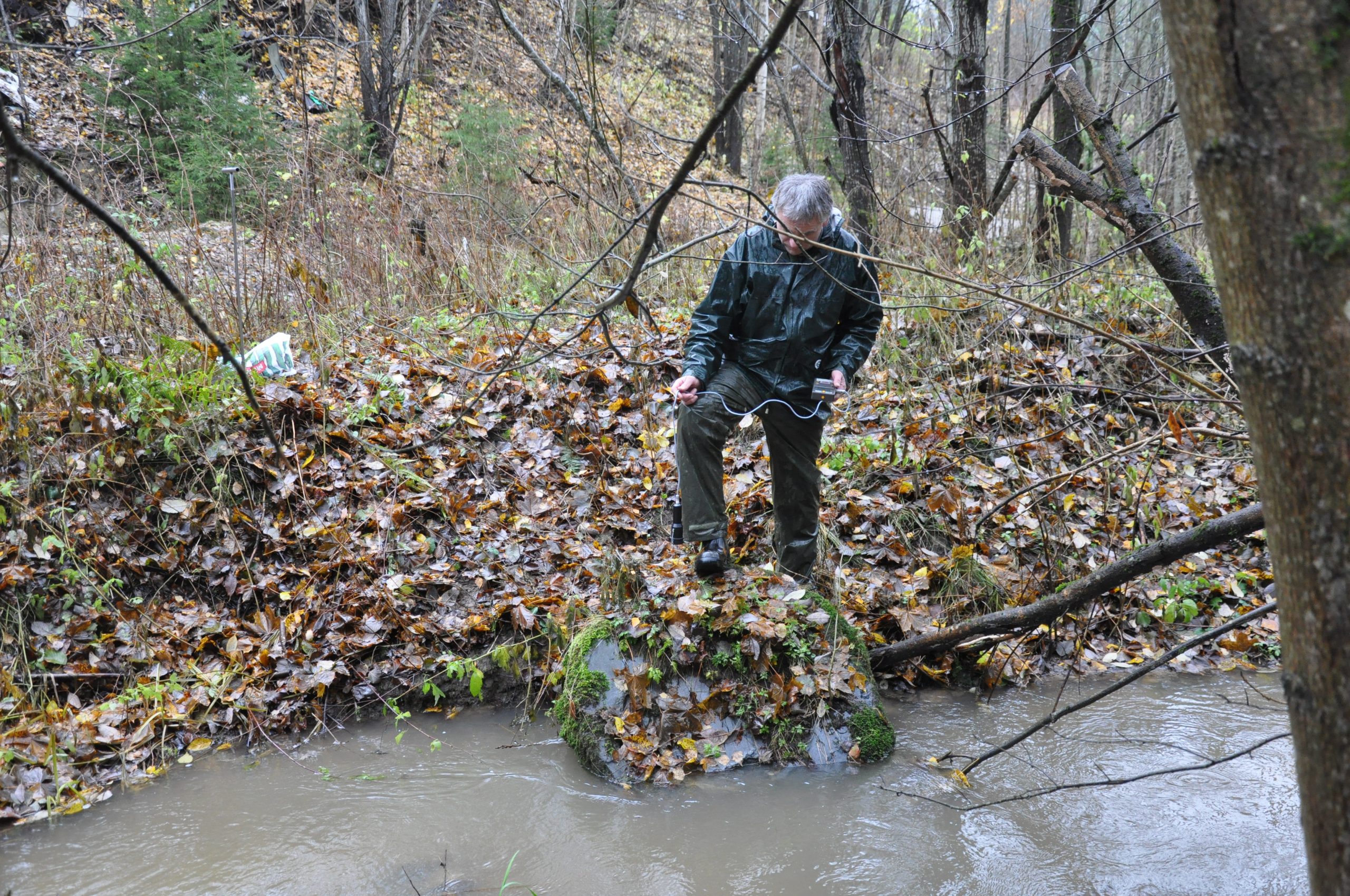 Bilde av en mann som står med foten på en stor slaggklump som ligger i ei elv. Mannen måler radioaktivitet.